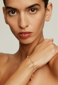 Pulsera de oro con cadena delicada, pequeñas cuentas blancas y un pequeño encanto. Lleva puesta en un brazo desnudo sobre un fondo neutro.