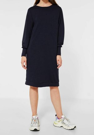 Kobieta ubrana w czarną sukienkę na długi rękaw o długości do kolan, zestawioną z białymi, masywnymi sneakersami z fioletowymi i żółtymi akcentami.