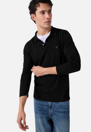Uomo che indossa una polo nera a maniche lunghe sopra una maglietta bianca, regolando la manica, abbinata a jeans blu, con volto leggermente rivolto a sinistra su sfondo bianco.