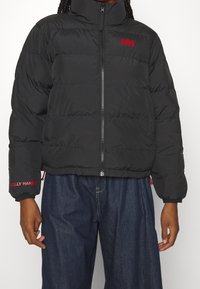 Zwarte pufferjack met een hoge kraag, voorzien van een rood Helly Hansen-logo. Het heeft een ritssluiting aan de voorkant en een lichtjes cropped pasvorm.