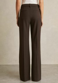 Persona che indossa pantaloni a gamba larga color marrone scuro a vita alta con tasche a filetto posteriori, in piedi contro una parete e un pavimento beige semplici.