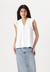 Top bianco in cotone con scollo a V e maniche a volant, caratterizzato da dettagli in pizzo e una vestibilità ampia. Abbinato a una gonna di jeans blu.