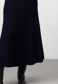 Marinblå stickad midikjol med en utflädd design, subtil textur och sidoflik. Bärs med knähöga svarta stövlar mot en neutral bakgrund.