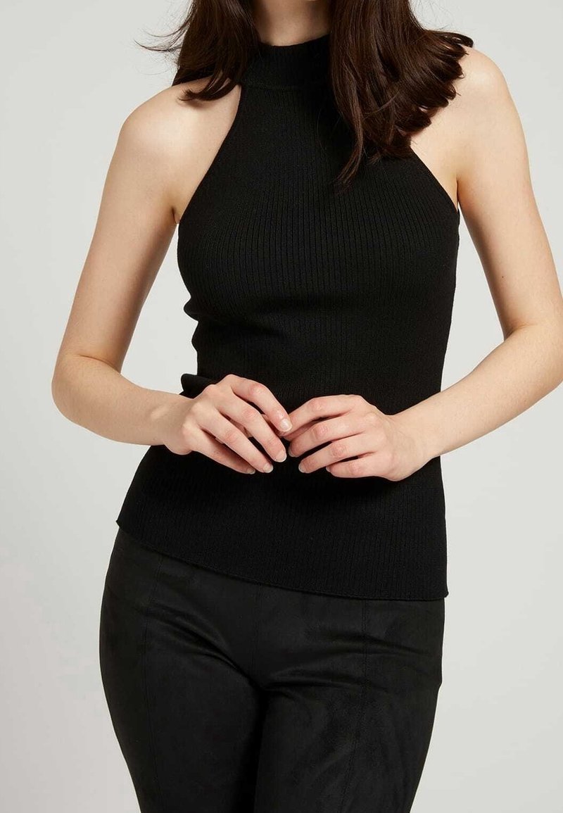 Femme portant un haut noir côtelé sans manches à col halter et un pantalon noir, les mains doucement jointes à la taille.