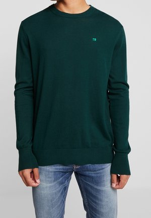 Maglione verde scuro a maniche lunghe con polsini e orlo a coste, indossato con jeans blu slavati, con un piccolo logo a contrasto sul petto.