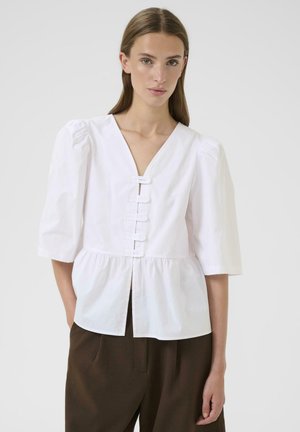 Blusa di cotone bianca con scollatura a V profonda, maniche a sbuffo e sei bottoni sul fronte. Presenta una vita arricciata con un orlo fluttuante.