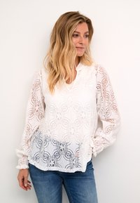 Blusa bianca in pizzo con motivi floreali, maniche lunghe a sbuffo e un sottile scollo a V. Abbinata a jeans blu, su uno sfondo neutro.