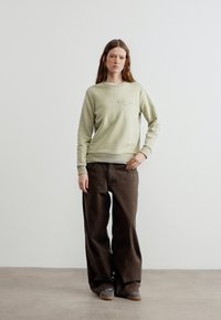 Γυναίκα που στέκεται με ουδέτερη έκφραση, φοράει ανοιχτό πράσινο φούτερ και φαρδιά καφέ παντελόνια μπροστά σε απλή λευκή τοιχοποιία.