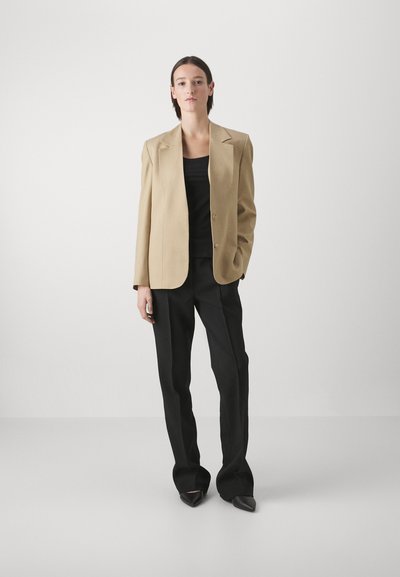 Beige taillierter Blazer mit breiten Revers, einreihiger Knopfverschluss, getragen über einem schwarzen Oberteil und kombiniert mit schwarzen Hosen, dazu spitze Absätze.