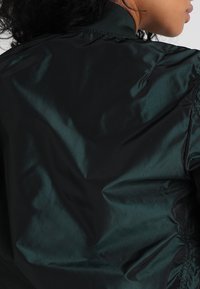 Czarna nylonowa kurtka bomberka o gładkiej, błyszczącej teksturze, z wysokim kołnierzem i elastycznymi mankietami, ukazująca eleganckie linie i minimalistyczny design.