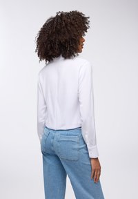 Blusa bianca a maniche lunghe con polsini a bottone, abbinata a jeans in denim azzurro chiaro a vita alta con tasche posteriori e una silhouette aderente.