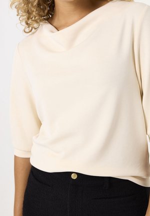 Light beige top featuring a cowl neckline and three-quarter sleeves, paired with black textured pants with a gold button detail.