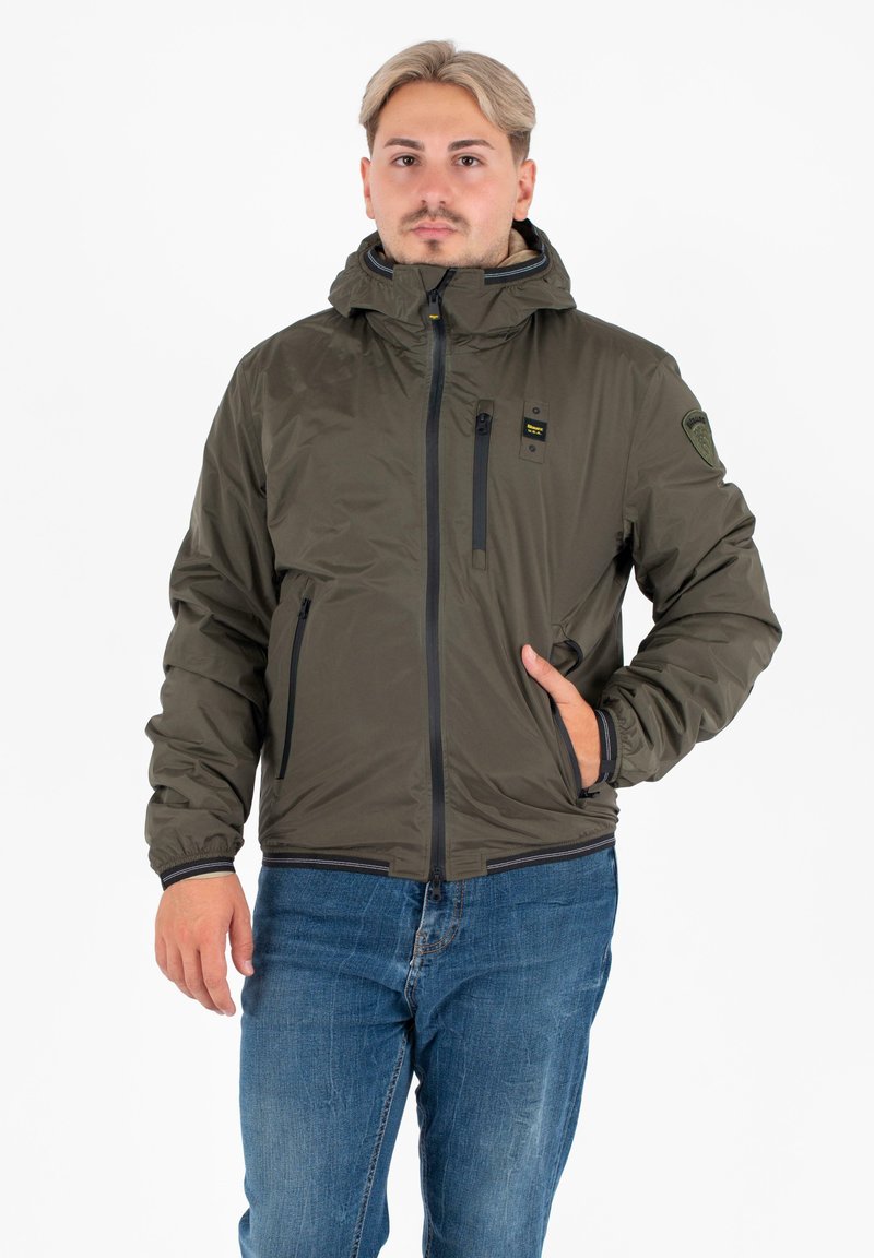 Giacca isolante verde oliva con cappuccio, zip frontale e tasche laterali. Presenta zip e dettagli neri, con polsini e orlo elasticizzati.