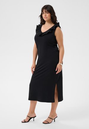 Femme portant une robe midi noire sans manches avec un col volanté et une fente latérale, associée à des sandales à talons noires à brides.