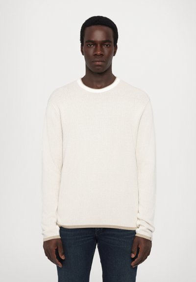 White ribbed long-sleeve shirt with a round neckline, featuring a textured surface and a relaxed fit, paired with dark blue jeans.