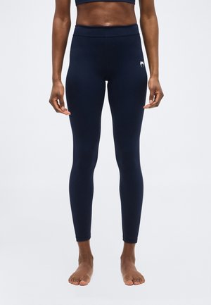 Személy tengerészkék magasderekú leggingset visel, bal combján egy kis fehér logóval, mezítláb áll egy sima fehér háttér előtt.