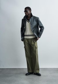 Mees, kes kannab musta nahktagi, kreemika värvi soonikkoes kampsunit, laiu oliivrohelisi pükse, musti kingi ja tumedaid päikeseprille, seisab halli tausta ees.