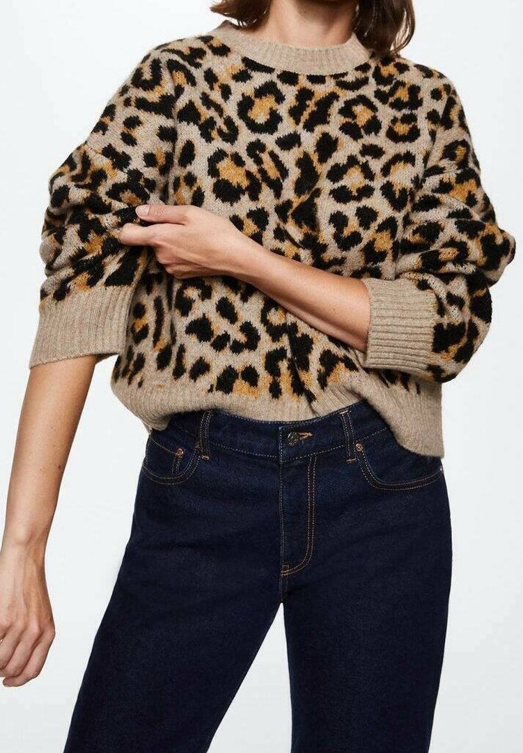 Femme portant un pull beige à imprimé léopard avec des poignets côtelés et un jean taille haute bleu foncé, bras croisés sur le corps ajustant le pull.