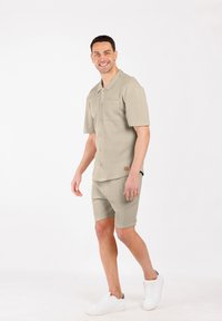 Homme souriant marchant, portant une chemise à manches courtes beige à boutons et un short assorti, avec des baskets blanches, sur fond blanc uni.