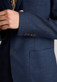 Blazer bleu marine en tissu texturé, avec deux poches avant, des poignets à trois boutons et un motif subtil avec un léger éclat.