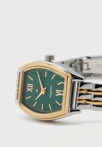 Ρολόι χειρός με πράσινο καντράν, χρυστούς ρωμαϊκούς αριθμούς, μεταλλικό λουρί σε χρυσό και ασημί και λογότυπο κορώνας πάνω από τα κεντρικά δείκτες που δείχνουν 9:55.