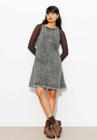 Robe en denim gris à la forme évasée, présentant un col dégagé et des coutures distinctes. Portée par-dessus un haut en dentelle noire à manches longues.