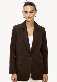 Blazer marron avec une fermeture à un seul bouton, des revers crantés, deux poches avant, et un tissu lisse et structuré. Porté sur un haut blanc.