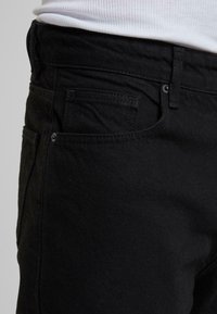Schwarze Jeans aus Denim mit glatter Textur, klassischem Schnitt, Vorder- und Gesäßtaschen sowie sichtbaren Nähten entlang der Säume.