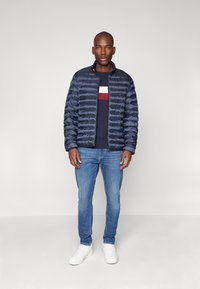 Mørkeblå pufferjakke med horisontale sømmer, mørkeblå genser med røde og hvite striper, lyseblå jeans og hvite sneakers.