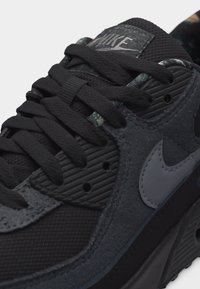 Κοντινό πλάνο μαύρου αθλητικού παπουτσιού Nike με σκούρες γκρι επενδύσεις από σουέντ, μαύρα κορδόνια και λογότυπο Nike στο πλάι και στη γλώσσα.