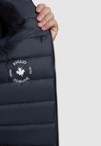 Tamsiai mėlynas pūkinis švarkas su siuvinėtu dizainu, su baltu logotipu ir tekstu kairėje pusėje šalia užtrauktuko. Matoma užtrauktuko kišenė.