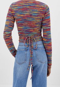 Achteraanzicht van een persoon die een meerkleurige gebreide omslagtrui draagt, vastgebonden op de taille, met blauwe high-waisted jeans met achterzakken.