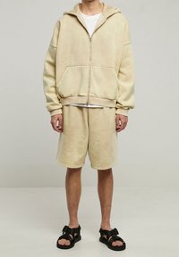 Felpa beige con zip e tasche frontali, abbinata a pantaloni corti, realizzata in tessuto morbido. Il modello indossa anche sandali neri. Design semplice e dalla vestibilità rilassata.