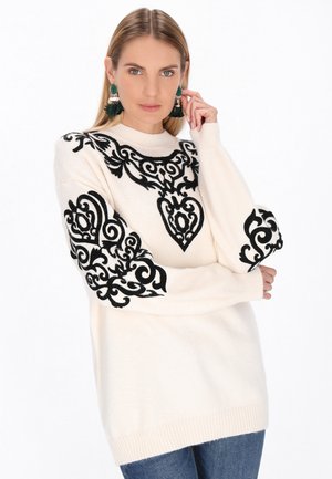 Creme sweater med intrikate sorte broderier på skuldrene og ærmerne, med ribbede manchetter og kant. Båret med blå jeans.