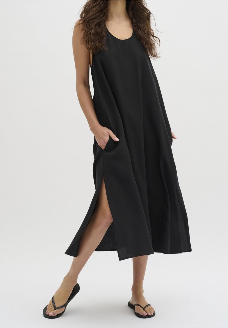 Femme portant une robe maxi noire sans manches avec des fentes latérales et des tongs noires, debout avec une main dans une poche.