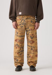Pantalones de pierna ancha beige con un estampado vintage en azul y rojo, que representa ilustraciones históricas y texto. Combinados con una sudadera marrón y zapatos negros.