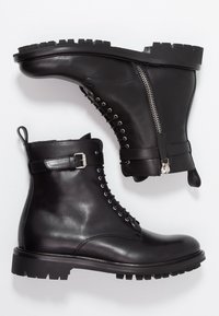 Botas de cuero negras hasta el tobillo con una suela robusta, con cremallera en la parte frontal, diseño de cordones y una correa lateral con detalle de hebilla.