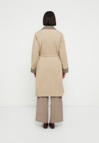 Trench-coat beige à taille ceinturée, avec un col à motif pied-de-poule et des poignets texturés, porté avec un pantalon beige à jambes larges.