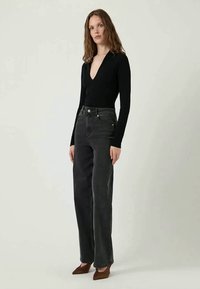Body nero a maniche lunghe con scollatura a V profonda, abbinato a jeans grigio scuro a vita alta. Scarpe marroni a punta completano il look.