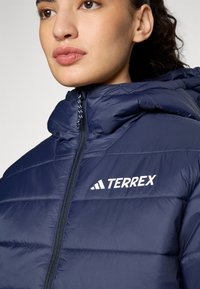 Marineblaue Steppjacke mit hohem Kragen, Reißverschluss und weißem "TERREX"-Logo. Glatte, glänzende Textur mit gesteppten Paneeldetails.