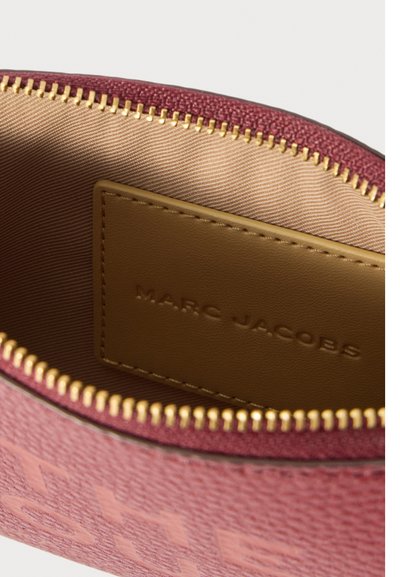 Rožinės Marc Jacobs pakuotės vidus, kuriame matyti smėlio spalvos odinė etiketė ir auksinė užtrauktuko užtrauktukas ant smėlio spalvos dryžuoto pamušalo.