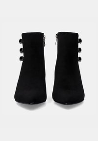 Botas negras de tobillo hechas de material suave, con una superficie lisa, forma de punta afilada y tres acentos brillantes a lo largo del costado.