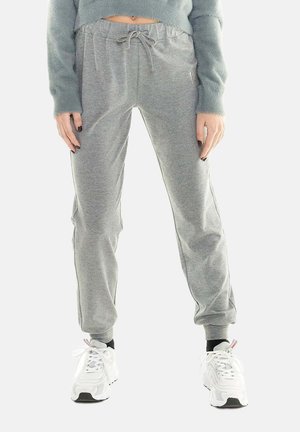 Femme portant un pantalon de survêtement gris à cordon, des baskets blanches, des chaussettes noires et un pull court gris duveteux, debout devant un fond uni.