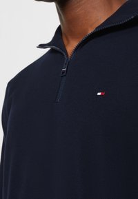 Pull-over bleu marine à demi-fermeture éclair en tissu lisse, avec un petit emblème rouge et blanc sur la poitrine et une fermeture éclair texturée.
