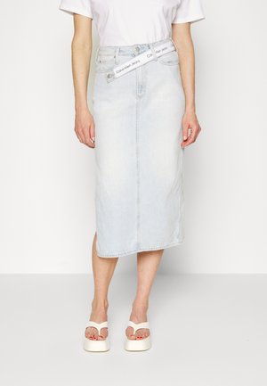 Osoba nosząca jasnoniebieską jeansową spódnicę midi z białym paskiem Calvin Klein Jeans, białe sandały i biały krótki top.