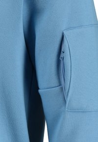 Sudadera de color azul claro con un tejido texturizado; presenta un pequeño bolsillo con cremallera en la manga izquierda para almacenamiento.