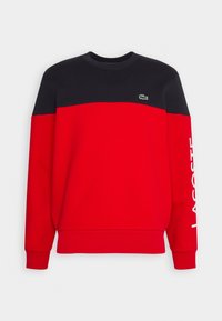 Svetřík v červené a námořnické modři se skládá z bavlněné látky, má kulatý výstřih a žebrované manžety. Na rukávu se nachází malé zelené logo a bílé nápis "LACOSTE".