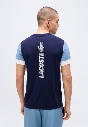 Uomo che indossa una camicia a maniche corte blu navy e azzurro chiaro con logo Lacoste bianco verticale e coccodrillo sulla schiena, di spalle.