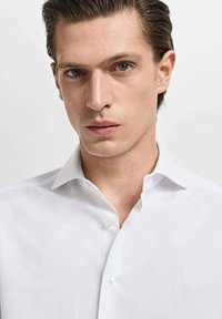 Camisa blanca de vestir con una textura suave, cuello puntiagudo y cierre frontal con botones. Presenta un acabado limpio y ajustado, sin patrones.