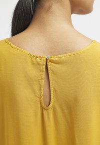 Haut jaune avec un col rond, un détail en forme de clé à l'arrière et une fermeture à bouton. Tissu lisse avec un drapé doux.